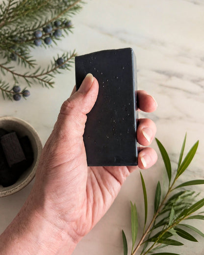 Hand holding a charcoal soap with ingredients on the background - Eternal Sunday