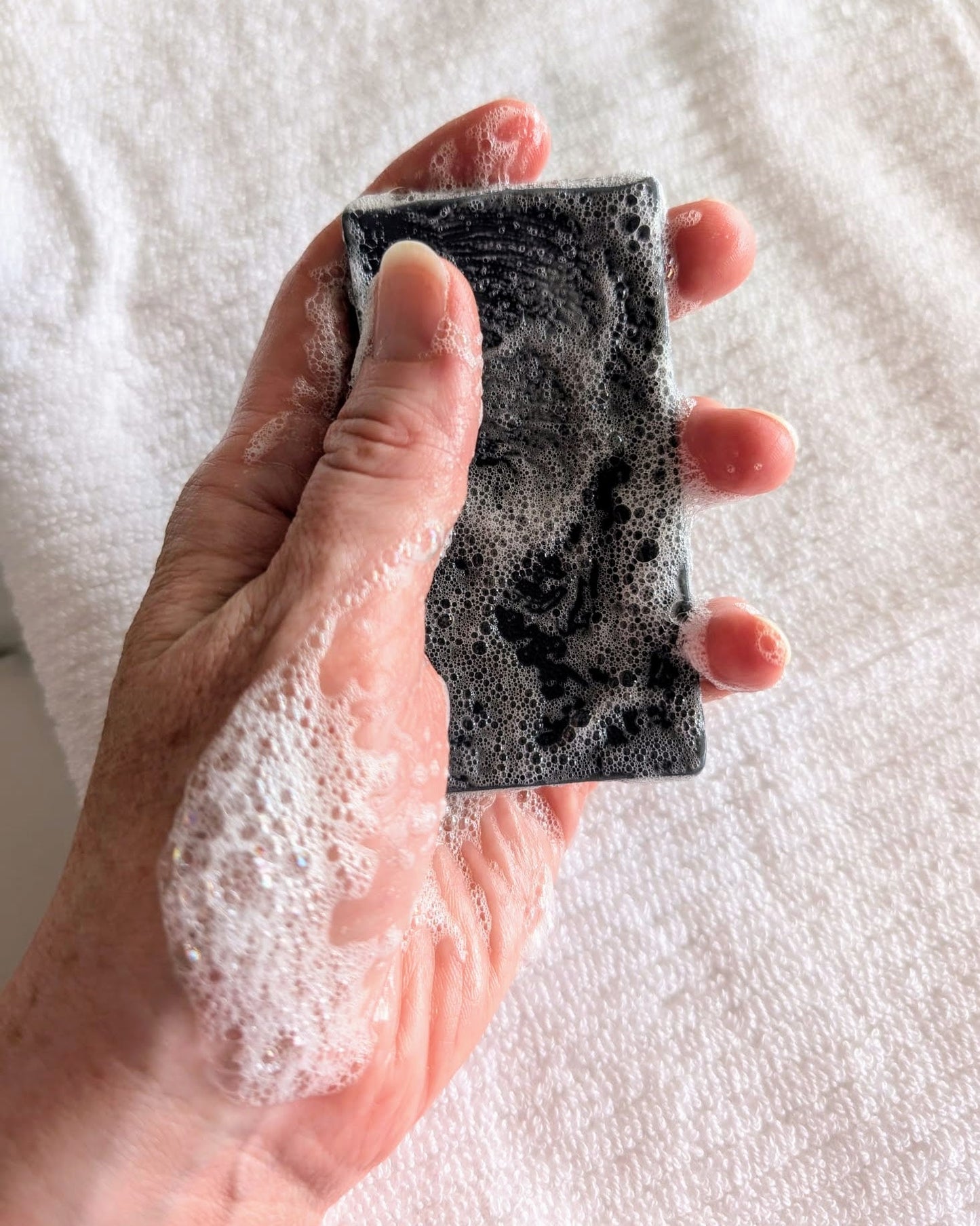 Hand holding the charcoal soap with a rich white lather - Eternal Sunday